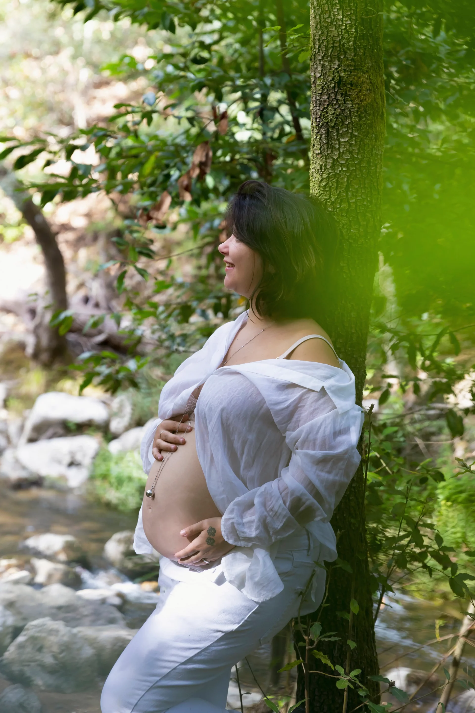 photo d'une future maman au bord d'une rivière, adossée à un arbre et enlaçant son ventre sandra-alvoet-photographe