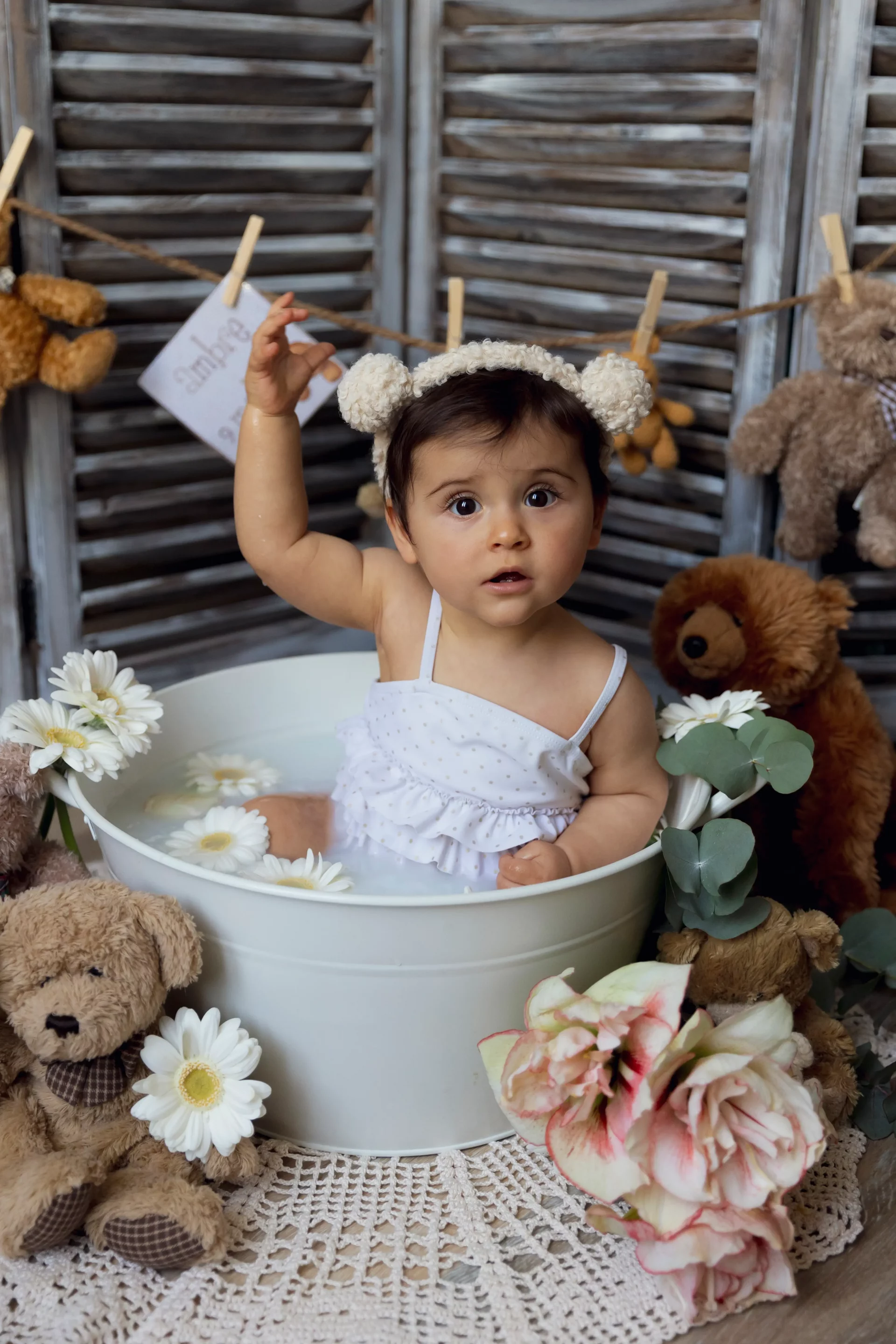 photographie-bain-de-lait bébé de 9 mois dans son bain de lait avec un maillot de bain blanc, entouré de fleurs et de nounours sandra-alvoet-photographe