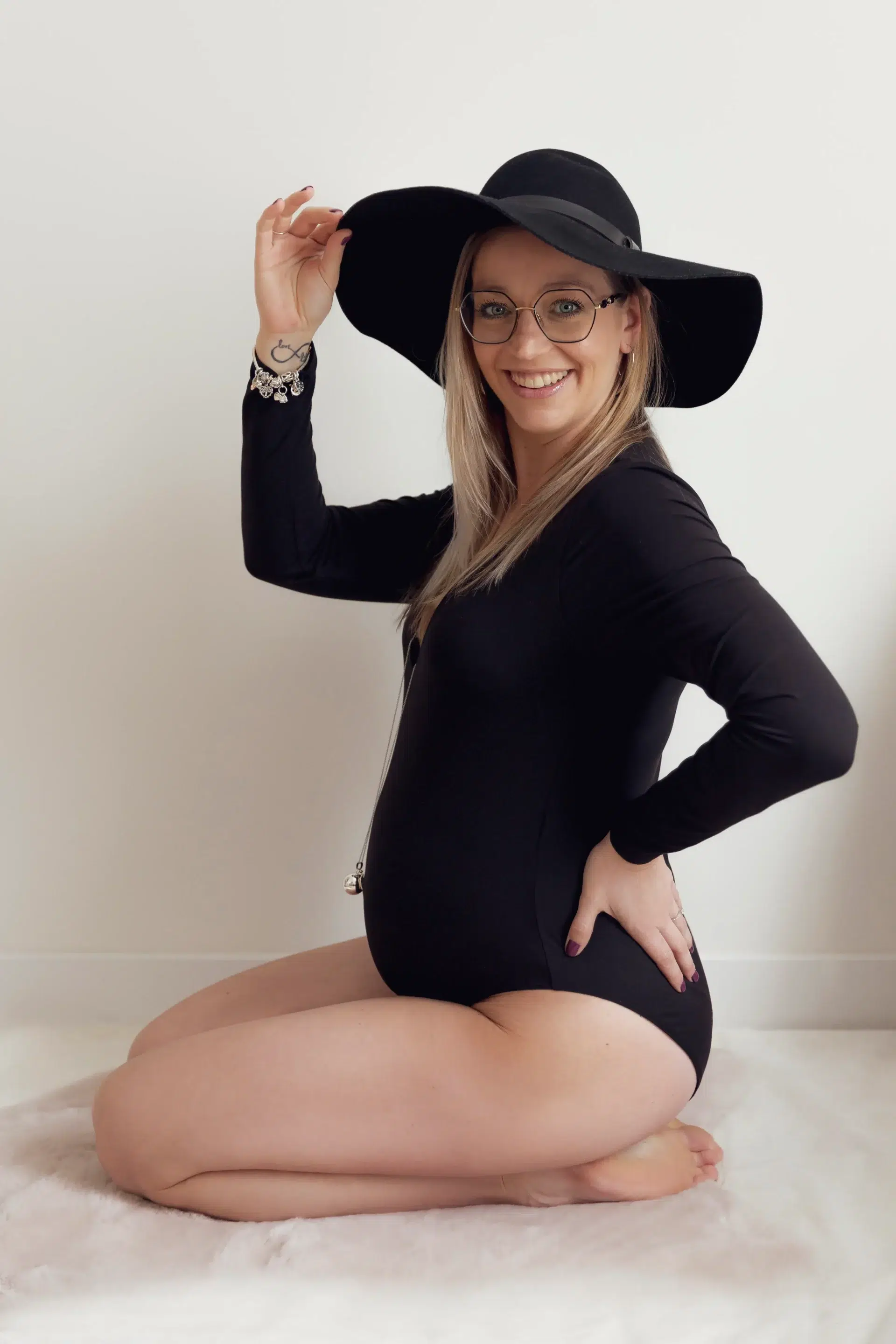 grossesse-nice Photographie d'une femme enceinte dans un studio photo à nice, vêtue d'un body et d'un chapeau noirs, souriante, et tenant le chapeau sandra-alvoet-photographe