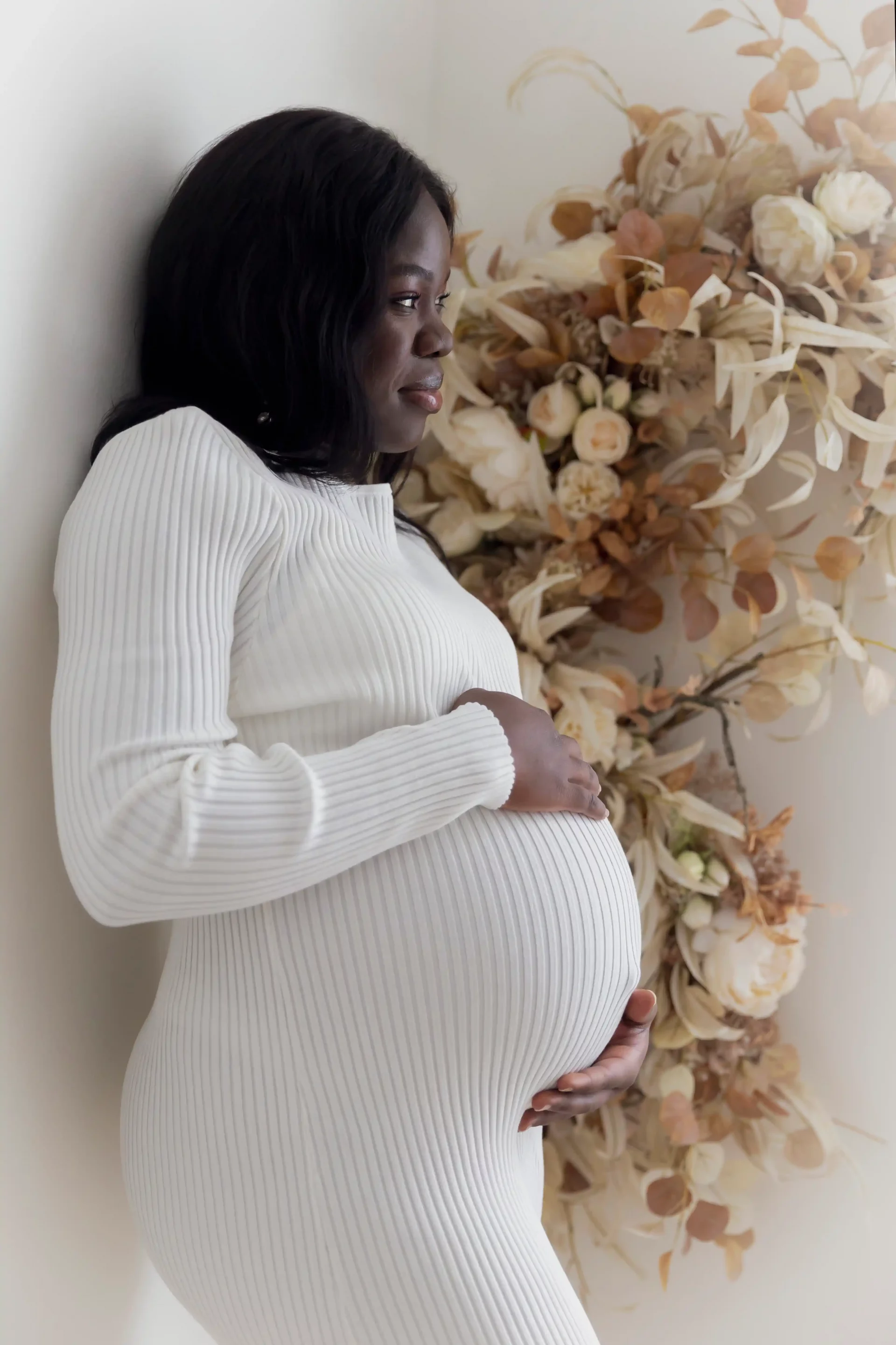 photo de grossesse d'une maman habillée en robe blanche, adossée à un mur de fleurs