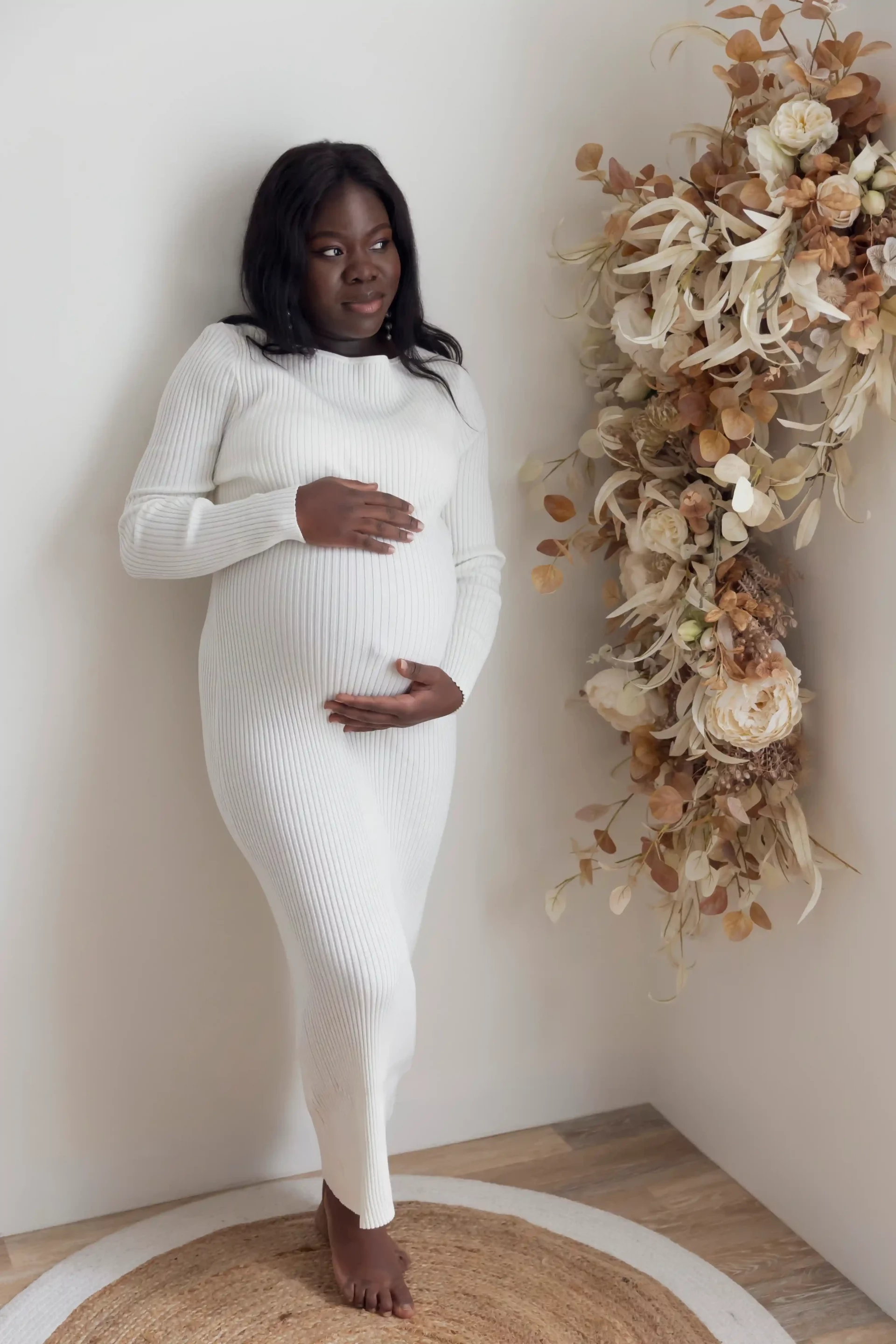 photo de grossesse d'une maman en robe blanche adossée au mur, à côté d'une guirlande de fleurs